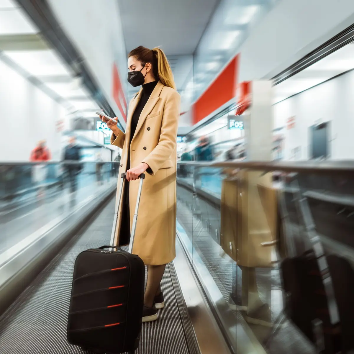 Airport traveler with carry-on luggage looking at her phone on a moving walkway