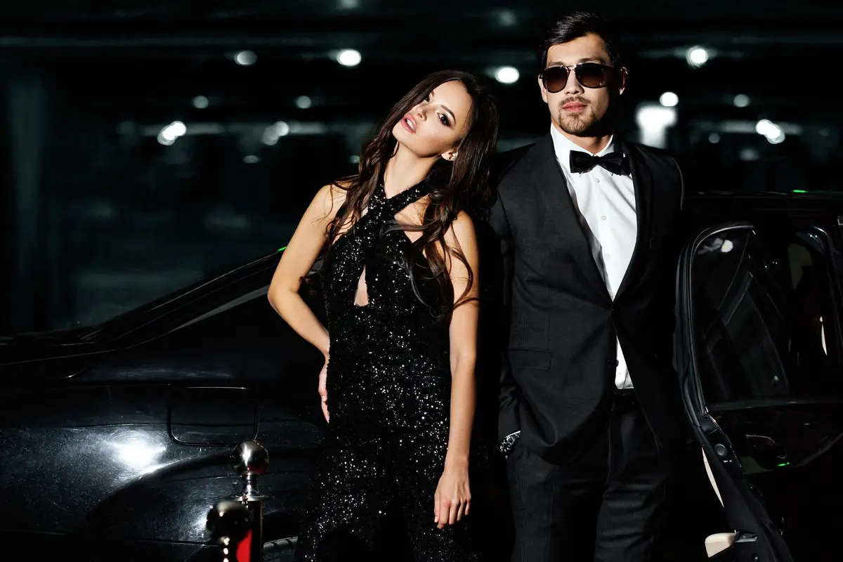 Couple in formal evening wear standing beside a black luxury car at night